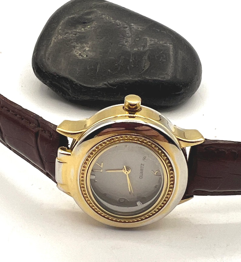 May include: A gold-toned quartz watch with a silver face and gold hands. The watch has a brown leather band. A dark gray stone is in the background.
