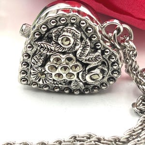 May include: Silver-toned heart-shaped pendant necklace with intricate floral and leaf detailing. The pendant features small, round embellishments and a small, silver chain. The background includes a red fabric.
