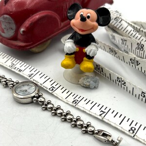 Op de afbeelding: Een rode speelgoedauto, een Mickey Mouse-beeldje, een zilveren horloge en een wit meetlint zijn gerangschikt op een wit oppervlak. Het horloge heeft een ronde wijzerplaat en een kralenband. Het meetlint toont centimeter metingen.
