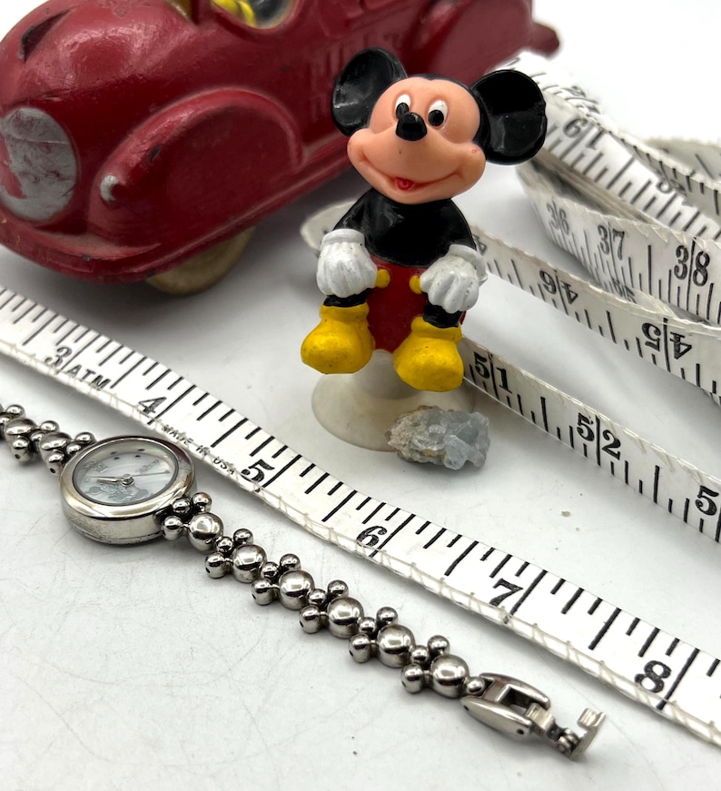 Op de afbeelding: Een rode speelgoedauto, een Mickey Mouse-beeldje, een zilveren horloge en een wit meetlint zijn gerangschikt op een wit oppervlak. Het horloge heeft een ronde wijzerplaat en een kralenband. Het meetlint toont centimeter metingen.