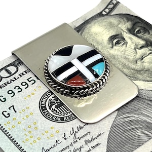 May include: A silver-toned metal money clip featuring a circular inlay with geometric patterns in black, white, turquoise, and reddish-brown. The inlay is bordered by a twisted silver-toned rope design. The clip is shown holding a 100 dollar bill.