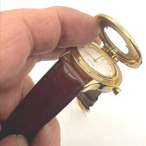 May include: A gold-tone quartz watch with a white face and brown leather band. The watch has a hinged cover that opens to reveal the watch face. The watch is held in a hand.