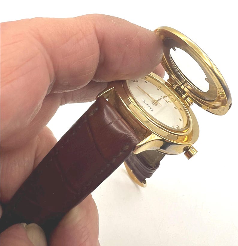 May include: A gold-tone quartz watch with a white face and brown leather band. The watch has a hinged cover that opens to reveal the watch face. The watch is held in a hand.