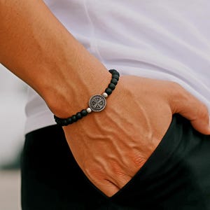 May include: Black beaded bracelet with a silver-colored circular centerpiece. The centerpiece features a cross design. The bracelet is worn on a wrist, with the hand resting in a pocket.