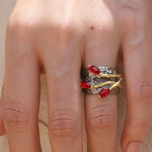 Puede incluir: Un anillo plateado con detalles dorados y piedras rojas. El anillo presenta una banda texturizada con ramas doradas y tres piedras rojas ovaladas. El anillo se lleva en un dedo.