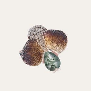Puede incluir: Anillo con forma floral con pétalos marrones texturizados, una sección de pequeños diamantes y una gema verde en forma de lágrima. El anillo tiene una banda plateada y un acento rosa claro.
