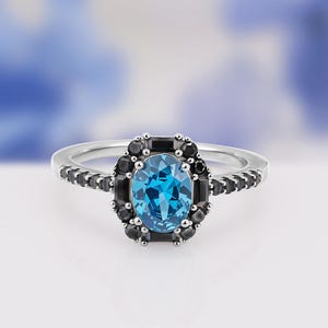 May include: A silver ring featuring a large oval, bright blue gemstone surrounded by a halo of small black stones. Additional black stones are set along the ring band, creating a striking contrast.