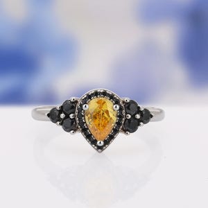 May include: A silver ring featuring a pear-shaped, yellow gemstone at its center, surrounded by a halo of black stones. Additional black stones are set on either side of the central design, creating a unique and elegant look.