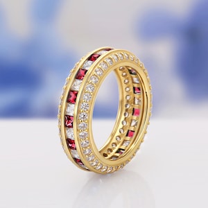 Könnte beinhalten: Ein goldener Ring mit abwechselnd roten und weißen quadratisch geschliffenen Steinen, die mit runden, klaren Steinen durchsetzt sind. Der Ring hat ein Doppelband-Design und ist vor einem weichen, verschwommenen Hintergrund platziert. Der Ring ist ein Schmuckstück.