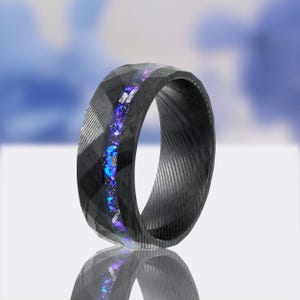 May include: A black ring with a geometric design and a central inlay of sparkling blue and purple stones. The ring has a textured surface and a polished finish, showcasing a modern and elegant aesthetic.