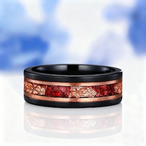 May include: A black ring with a copper-colored band and a center filled with red and gold-colored stones. The ring is set against a blurred blue and white background.