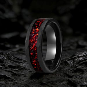 Può includere: Un anello nero con una texture martellata e un vivace centro rosso. La fascia dell'anello è nera lucida e il centro presenta un design rosso maculato e infuocato. L'anello è un primo piano su uno sfondo scuro.