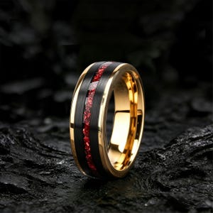 Op de afbeelding: Een ring met een geborstelde zwarte middenband met een dunne, verzonken inleg van vermalen rode steen. De randen van de ring zijn van gepolijst goud en het binnenoppervlak is ook afgewerkt in een gladde, reflecterende goudtint.