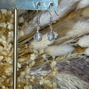 May include: A pair of clear crystal earrings with silver hooks. The earrings feature round, textured crystal beads. They are displayed on a silver metal stand, set against a blurred backdrop of dried flowers. The earrings are a delicate accessory.
