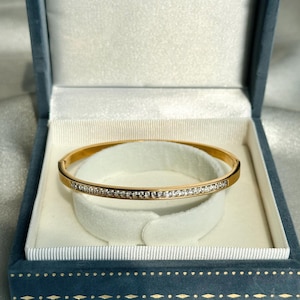 以下が含まれることがあります： 小さな透明な宝石が並んだゴールドカラーのバングルブレスレット。ブレスレットは、白いサテンの内装が施された青いベルベットのジュエリーボックスの中にあります。