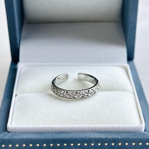 May include: Open-ended silver ring with a floral design, presented in a blue jewelry box with a white interior. The ring features a detailed pattern of small, sparkling stones.