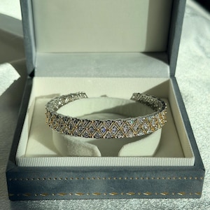 May include: A silver and gold bracelet with a diamond pattern is displayed inside a gray jewelry box. The bracelet is a cuff style, with a geometric design of diamonds and gold accents. The box has a soft, velvet-like interior.