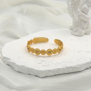 May include: A gold-toned open cuff bracelet featuring a repeating pattern of small, textured daisy-like flowers. The band has a delicate, intricate design with a central row of floral motifs and a perforated, dotted texture along the inner edges.