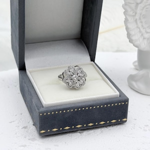 May include: A silver-toned ring featuring a flower-shaped cluster of baguette and round-cut clear stones, presented inside a dark grey velvet-lined jewelry box with gold-colored decorative trim along the exterior edge of the base.