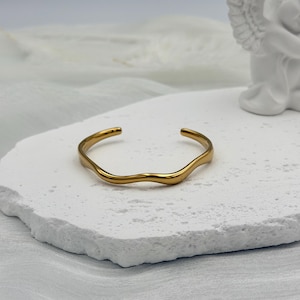 Puede incluir: Un brazalete de metal en tono dorado que presenta un diseño ondulado continuo y suave. La pulsera tiene una estructura abierta que permite su ajuste y posee un acabado de superficie pulido y reflectante en toda la pieza.