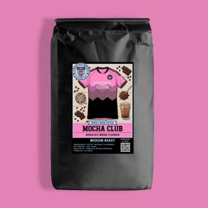 May include: A black bag of whole bean coffee with a label featuring a pink and black soccer jersey design. The label reads "MOCHA CLUB" and "CHOCOLATE MOCHA FLAVORED". The bag is on a pink background.