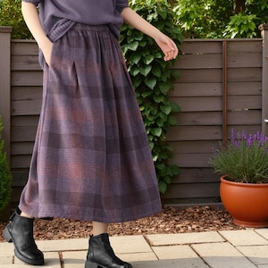 May include: A long, purple and red plaid skirt with a matching top. The skirt features a pleated design and pockets. The outfit is completed with black ankle boots. The background includes a wooden fence and greenery.