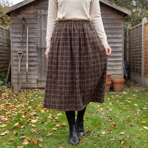 May include: A midi-length skirt featuring a brown and black plaid pattern with a subtle textured weave. The skirt has an elasticated waistband and a full, gathered silhouette that falls below the knees, paired with a cream long-sleeve top.