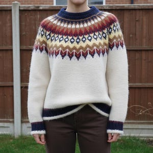 Puede incluir: Suéter color crema con canesú estampado en tonos rojos, amarillos y marrones. El suéter tiene cuello y puños azul marino con rayas rojas. Una prenda cálida y acogedora para el clima frío.