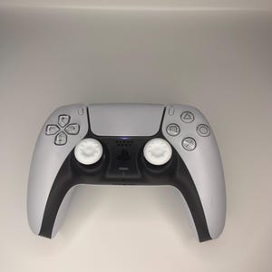 Puede incluir: Un mando de videojuegos inalámbrico blanco y negro con una sección central negra y botones blancos. El mando cuenta con botones direccionales, botones de acción y dos palancas analógicas. El diseño es elegante y moderno, adecuado para jugar.