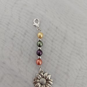 Könnte beinhalten: Ein silberfarbener Charm mit einem Sonnenblumenanhänger und einer Reihe farbiger Perlen. Die Perlen sind in Gold-, Grün-, Lila- und Koralltönen gehalten. Der Charm hat einen Karabinerverschluss zum einfachen Anbringen.