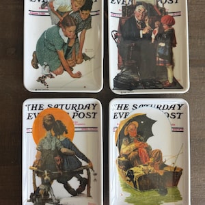 Puede incluir: Un juego de cuatro bandejas rectangulares de cerámica blanca, cada una con una ilustración diferente de una portada de la revista The Saturday Evening Post. Los diseños muestran diversas escenas con niños y adultos.