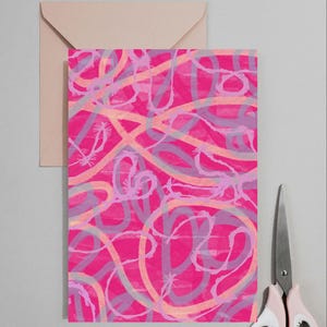 Puede incluir: Una tarjeta de felicitación rectangular con un fondo rosa intenso y bucles y líneas abstractas superpuestas en tonos morado claro y melocotón suave. La tarjeta está colocada frente a un sobre rosa claro y junto a unas tijeras de metal.