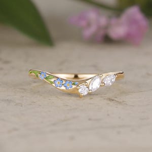 May include: A gold curved band ring featuring a series of small blue enamel forget-me-not flowers with green leaves on one side and three clear faceted stones, including one round and two marquise-shaped gems, on the other side of the band.