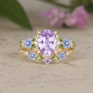 May include: A gold ring set featuring a central oval-cut light purple gemstone. The band is decorated with small blue enamel flowers, green enamel leaves, and tiny clear accent stones. A matching curved band sits below with similar floral details.