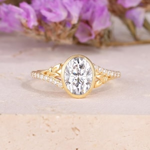 Puede incluir: Un anillo de oro con una piedra preciosa clara de corte ovalado como pieza central. La banda presenta pequeñas piedras brillantes y diseños en forma de hojas en los laterales. El anillo se encuentra sobre un fondo suave y neutro, resaltando su elegante diseño.