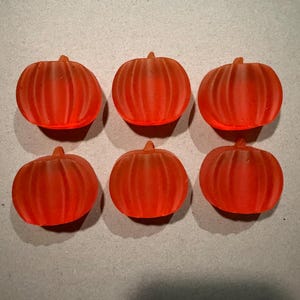 May include: Six small, translucent orange pumpkin-shaped objects arranged in two rows of three. Each pumpkin features a smooth, frosted texture with vertical ridges and a small stem at the top, creating a uniform appearance across all pieces.