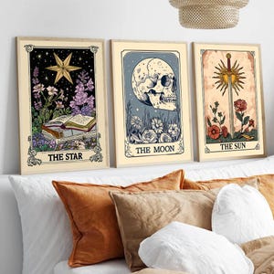 Könnte beinhalten: Drei gerahmte Tarotkarten-Drucke. Die Sternkarte zeigt einen goldenen Stern, Blumen und ein offenes Buch. Die Mondkarte zeigt einen Schädel mit Blumen. Die Sonnenkarte zeigt ein Schwert und eine Sonne mit Blumen. Alle Karten haben Text.