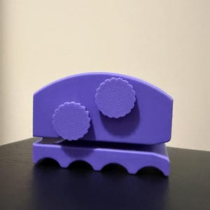 May include: A purple, handheld paper craft tool with a scalloped edge design. The tool has two circular, textured knobs on top and a curved top. The base has a series of curved cutouts. The tool is sitting on a dark surface.