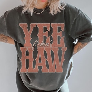 May include: Black trucker hat with "DEAD LAST RACING" text and a skull graphic. A dark gray oversized t-shirt features the text "YEE f*ckin HAW" in a distressed, reddish-brown font. The outfit is completed with black leggings.