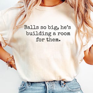 May include: Cream-colored t-shirt with the text "Balls so big, he's building a room for them" in black. The shirt is a crew neck style and is paired with blue denim jeans. The model is wearing a gold necklace and bracelets.