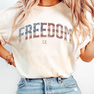 May include: Cream-colored t-shirt with the word "FREEDOM" in distressed red, white, and blue lettering. The initials "C.K." are printed below the word. The shirt is paired with blue denim jeans.