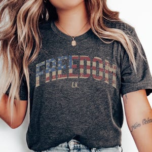 May include: Dark gray t-shirt with the word "FREEDOM" in a distressed, patriotic design. The letters are filled with a pattern of the American flag. Below the word "FREEDOM" is the text "C.K."