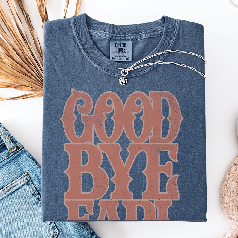 K&ouml;nnte beinhalten: Ein marineblaues T-Shirt mit dem Text "GOOD BYE LADY" in einem verblichenen, orangefarbenen Schriftzug. Das Shirt hat einen Rundhalsausschnitt und kurze &Auml;rmel. Eine silberne Halskette mit einem Sonnenblumenanh&auml;nger liegt auf dem Shirt.