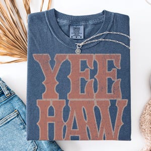 Pu&ograve; includere: Una maglietta blu navy con la scritta "YEE HAW" in un carattere rosso-arancio sbiadito. La maglietta ha un girocollo e maniche corte. Una collana d'argento con un ciondolo circolare &egrave; drappeggiata sulla maglietta. La maglietta &egrave; piegata su una superficie bianca.