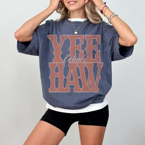 May include: A blue-grey oversized t-shirt with the text "YEE f'ckin HAW" in a brown, western-style font. The shirt has a white undershirt peeking out at the neckline and hem. The model is wearing black shorts.