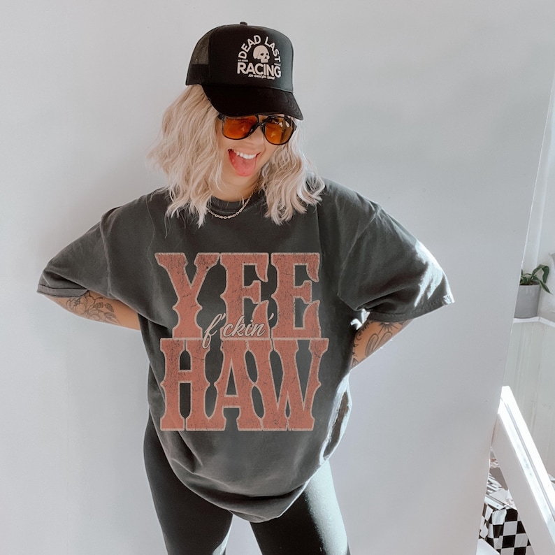 May include: Black trucker hat with "DEAD LAST RACING" text and a skull graphic. A dark gray oversized t-shirt features the text "YEE f*ckin HAW" in a distressed, reddish-brown font. The outfit is completed with black leggings.
