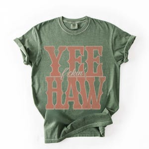 May include: Olive green t-shirt with the words "YEE f*ckin HAW" in a distressed, western-style font. The text is in a reddish-brown color, contrasting with the shirt's base color. The shirt has a crew neck and short sleeves.