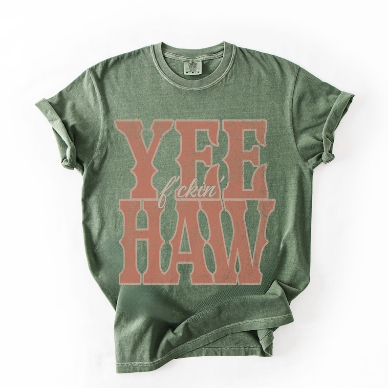 May include: Olive green t-shirt with the words "YEE f*ckin HAW" in a distressed, western-style font. The text is in a reddish-brown color, contrasting with the shirt's base color. The shirt has a crew neck and short sleeves.