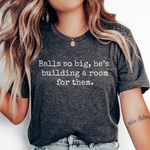 May include: Dark gray t-shirt with the text "Balls so big, he's building a room for them" in white. The shirt is a crew neck style and appears to be made of a soft material. The shirt is being worn.