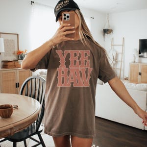 Pu&ograve; includere: Una maglietta marrone con la scritta "YEE HAW" in un carattere color corallo in stile vintage. La maglietta &egrave; di vestibilit&agrave; ampia. Viene indossato un berretto da baseball nero con testo bianco.